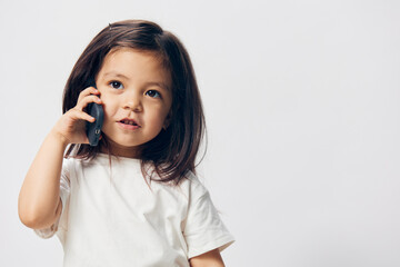 a little girl of preschool age, sitting on a white background in a white T-shirt, enthusiastically talking on the phone, turning half-side to the camera. The topic of the conversation on the phone