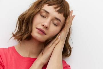 a gentle, relaxed, sleepy woman stands in a red T-shirt on a white background and holds her hands folded in her palms under her head, closing her eyes