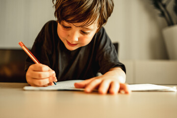 Naklejka na ściany i meble A five - year - old boy is doing his homework . A boy with a pencil in his hands writes, a boy draws on white paper at the table. Primary school and home schooling, the concept of distance education.