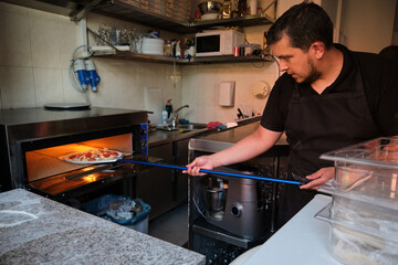 Latin man putting pepperoni pizza into the oven in a restaurant kitchen. Pizzeria.