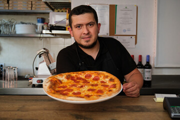 Latin male chef holding a freshly baked pepperoni pizza. Pizzeria. - obrazy, fototapety, plakaty