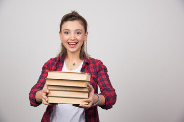 A happy woman carrying a stack of books on a gray wall