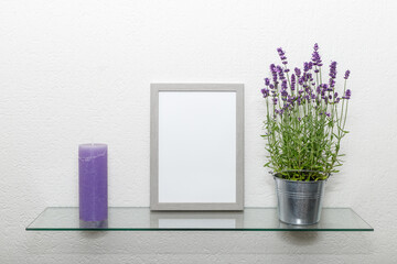 Blank grey wooden photo frame, lavender flowers in zinс pot, violet vintage candle on glass shelf...