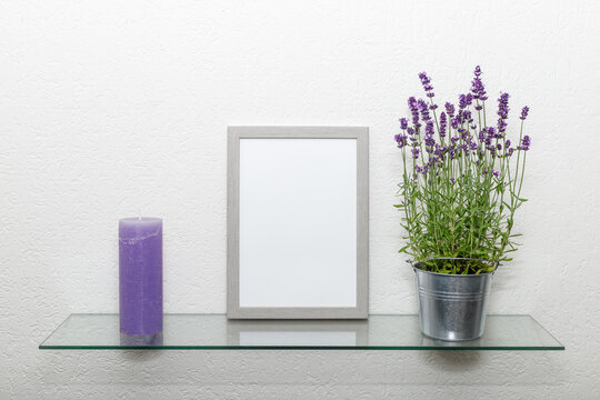 Blank grey wooden photo frame, lavender flowers in zinс pot, violet vintage candle on glass shelf against light wall.
