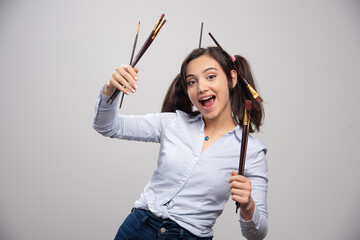 Young woman artist with paintbrushes smiling on gray background