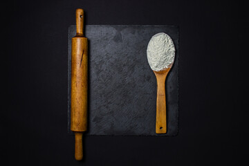 Wooden spoon with flour and a rolling pin on a black background. The concept of baking and confectionery.