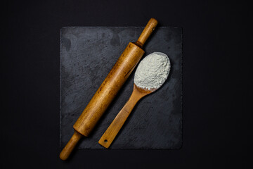Wooden spoon with flour and a rolling pin on a black background. The concept of baking and confectionery.