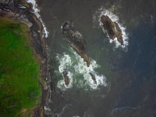 View from above. Large stones, boulders in the water. Green hilly coast. White foamy waves. Silence and serenity. Calm scenes. Leisure, romance, adventure, ecology, geology.