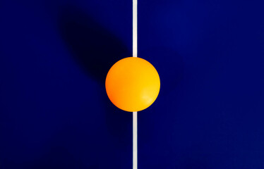 An orange ping pong ball flat lay at the center of the white line on dark blue table tennis court with natural sunlight, high angle view. Draw game idea. Concept of symmetry, abstract.