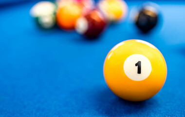A number 1 yellow billiard at a blue pool table surface with blurred background of colorful balls. Indoor sport and recreation, competitive and winner achieve concept. Game challenge.