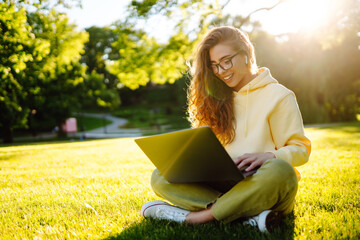 Young woman with wireless headphones working online or studying sitting on grass at park. Business, blogging, freelance, education concept.