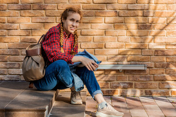 A smiling woman with pigtails in a plaid shirt and jeans sits on the steps against the backdrop of a brick wall. Street fashion.