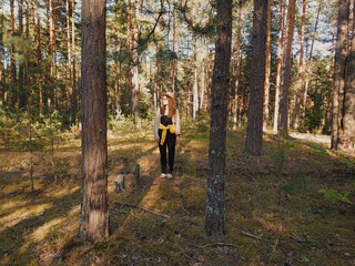 Fototapeta premium Young woman standing in the evergreen forest on a sunny summer day. Glasses, hat, denim overalls, sneakers. Mighty pine trees. Nature, hiking, trekking, ecotourism themes. Russia, Russian nature