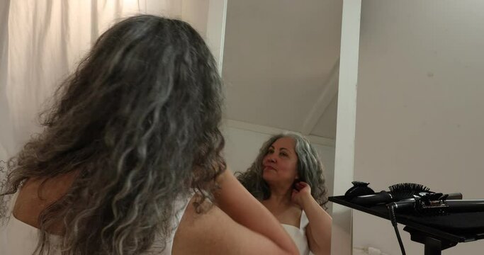 Mature woman looking at herself in front of the mirror, untying her long gray-black wavy hair, running her hand through her hair, reflected in the mirror with loose hair, smiling on white background