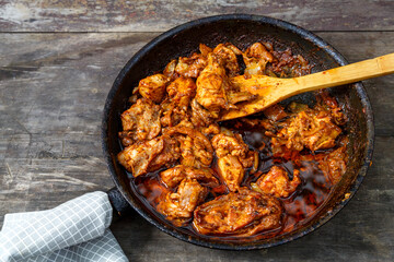 Fried chicken with spices in a frying pan with a wooden spatula on a dark table.