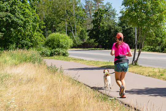 A female athlete in a pink T-shirt, shorts and a red baseball cap runs with a dog along the track. Canicross. Sports running with a dog. Lifestyle.