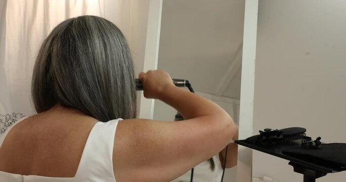 Woman giving the final touch to her gray-black hair that she straightened herself, combing it with a comb and sliding the straightening iron over the hair, reflected in the mirror, white background
