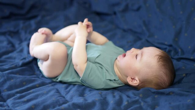 Lovely Caucasian baby boy lies on bed with bare feet. Cute kid rising legs up and mumbling something.