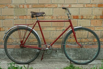 a very old men's cherry-coloured iron road bike with chrome fenders stands on the street at daytime near a brick wall in summer