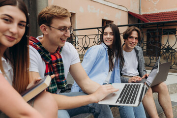 Group of cheerful students teenagers in casual outfits with note books and laptop are studying outdoors