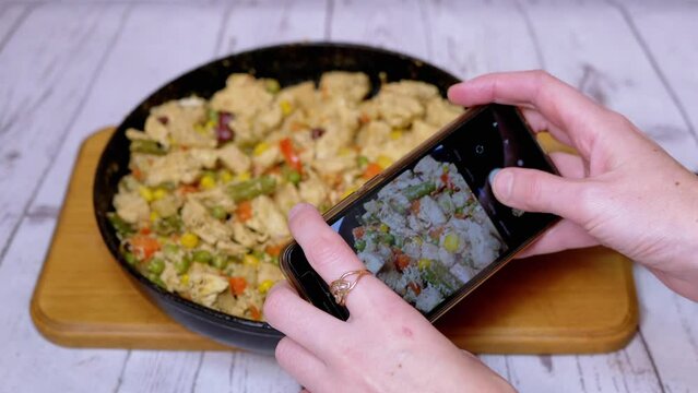 Female Taking Photo of Fried Meat with Vegetables on a Smartphone Camera. Close-up of a Frying Pan with juicy stewed chicken fillet pieces, mixed with colorful vegetables. Home kitchen. Blogging.