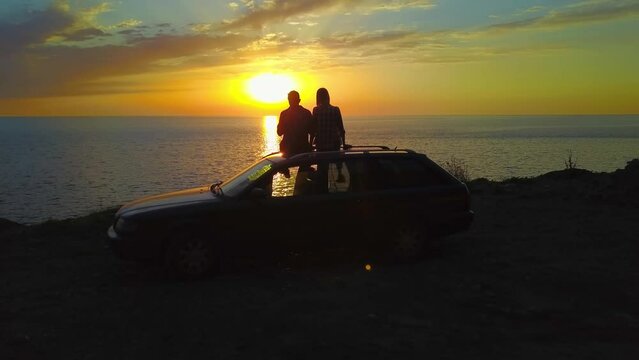 Man and a woman are sitting on the roof of a car on a cliff in front of the sea with a charming beautiful orange-blue sky at sunset, a view from a drone. The concept of rest and travel, love, romance