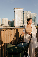 Full-length portrait of stylish young mother in summer light outfit walking in city with baby carriage and enjoying summer. Woman with baby in city background 