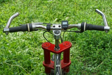 Fototapeta na wymiar a metal handlebar with black rubber grips and a thick cherry-colored iron fork from a mountain sports bike against the background of green grass on the street in summer