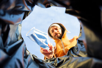 Man standing outside in sunshine wearing a brown hoodie throwing a covid surgical mask into a trash...