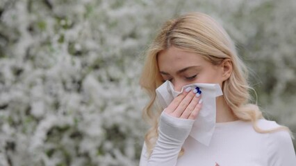 Sick displeased female with watery eyes blowing runny nose in paper tissue during walk in park. Unhappy beautiful lady suffering from seasonal allergy outdoors.