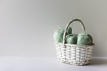 Fototapeta na wymiar Light green yarn threads in a white basket on a gray background with knitting needles.