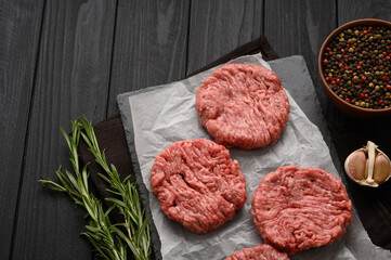Raw beef burger patties on butcher's wooden board, rosemary and pepper. Black background. View from above.