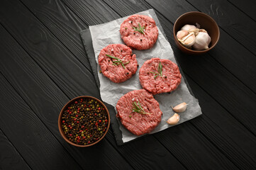 Raw beef burger patties on butcher's wooden board, rosemary and pepper. Black background. View from above.