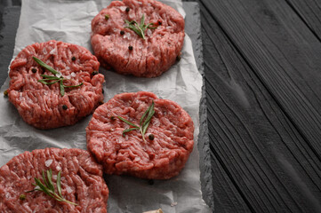 Raw beef burger patties on butcher's wooden board, rosemary and pepper. Black background. View from above.