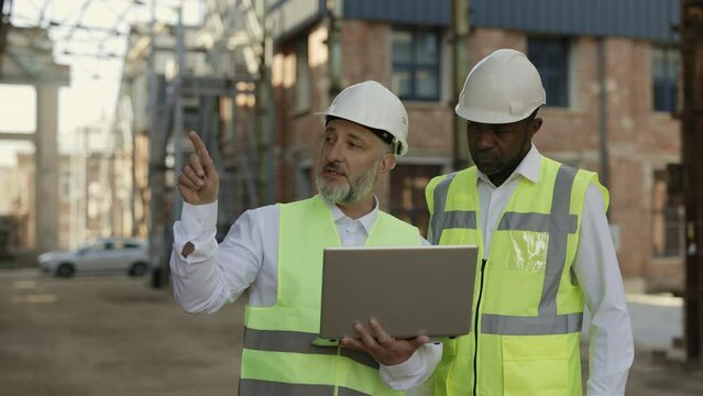 Caucasian aged architect holding portable laptop and explaining ideas to africa american construction worker. Multiethnic partners in protective uniform discussing structural process at building area.
