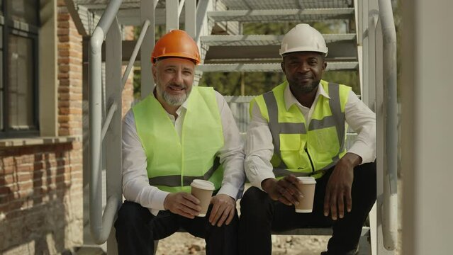 Smiling multicultural builders in hardhats looking at camera with smile while sitting together on stairs at construction site. Colleagues talking during enjoyable coffee break.