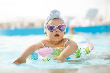 Cute funny toddler girl in colorful swimsuit and sunglasses relaxing on inflatable toy ring floating in pool have fun during summer vacation in tropical resort. Child having fun in swimming pool. 