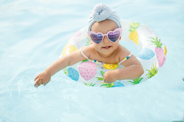 Cute funny toddler girl in colorful swimsuit and sunglasses relaxing on inflatable toy ring floating in pool have fun during summer vacation in tropical resort. Child having fun in swimming pool. 