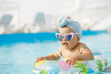 Cute funny toddler girl in colorful swimsuit and sunglasses relaxing on inflatable toy ring floating in pool have fun during summer vacation in tropical resort. Child having fun in swimming pool. 