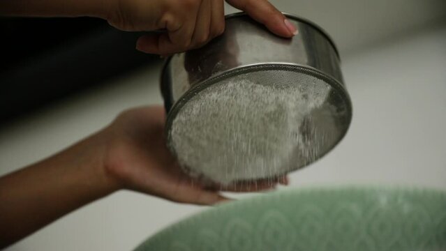 Cooking in the kitchen. Closeup view of female hands knocking a metallic sifter to sift flour into a bowl.
