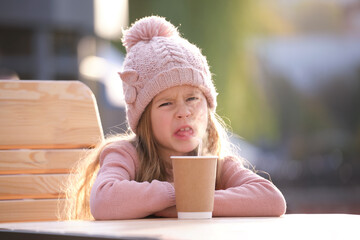 Portrait of cute little child girl in pink hat sitting alone at street cafe drinking tea from paper cup