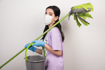 Woman with medical facemask posing with broom and bucket on white background