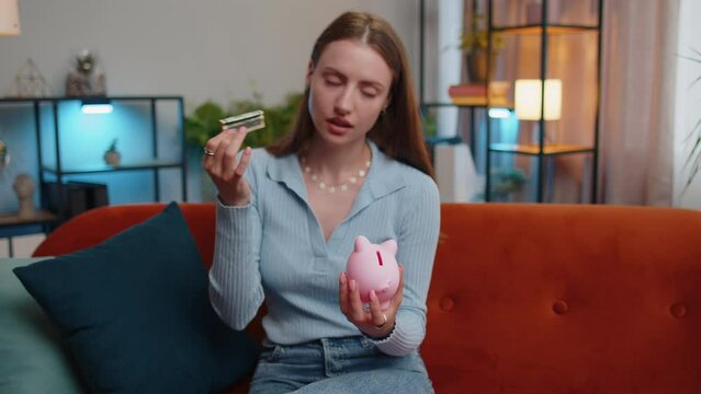 Poor young woman insufficient amount of money, holding piggybank and one dollar banknote at home. Financial crisis. Bankruptcy. Poverty and destitution. Depressed sad girl in living room on couch