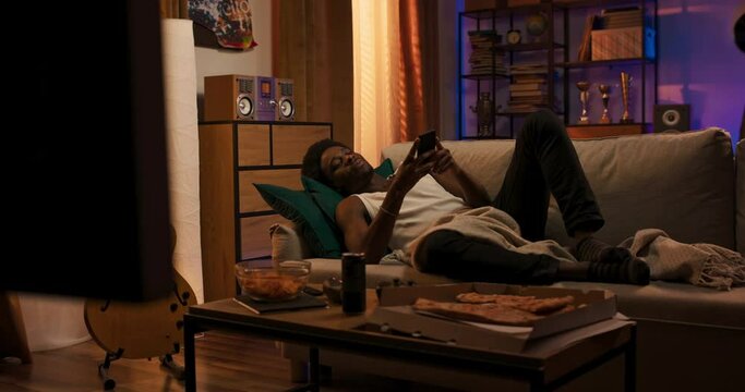 Young man holds smartphone texting or playing mobile games lying on couch at home in evening using social media app to send messages browsing websites on phone pizza on coffee table next to him.