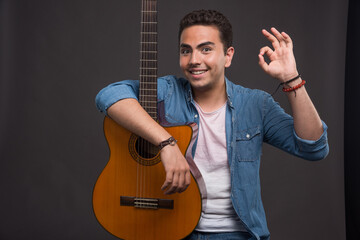 Happy man holding a guitar and making ok sign on dark background