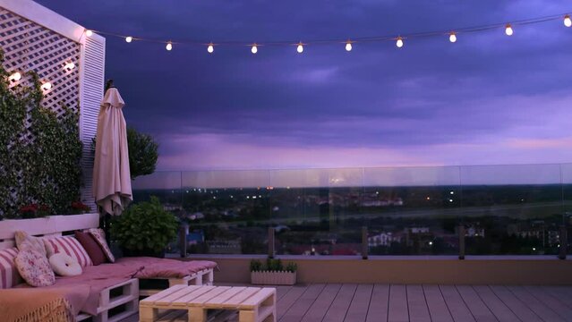 rooftop patio garden with light strings, glass balustrade and beautiful view on evening city in summer twilight