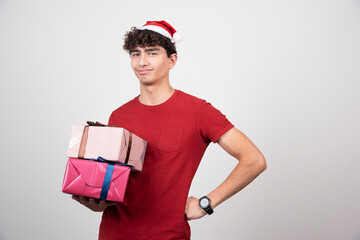 Serious male in Santa hat holding gifts on gray background