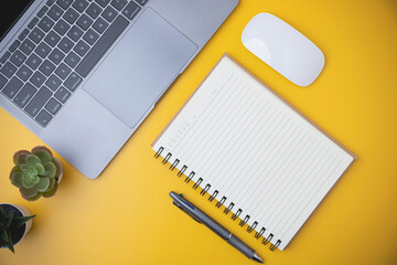 Business, technology, study, and learning concept. Flat lay of office yellow desk with laptop, and a white mouse. Top view blank notebook with a pen on yellow table with copy space.