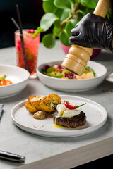 beef steak, beef patty with poached egg and mini potatoes on white plate, pepper falls from the mill, frozen photo, vertical