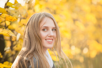 Portrait of a beautiful young woman against a background of colorful golden foliage in an autumn park. Attractive woman with light brown hair near bushes with yellow leaves. Fall season. Close-up.